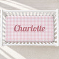 Top-down view of a white wooden crib containing a pink and white gingham crib sheet with the name 'Charlotte' printed in dusty pink script across the center.