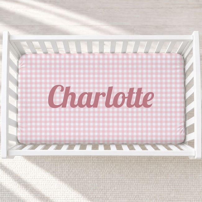 Top-down view of a white wooden crib containing a pink and white gingham crib sheet with the name 'Charlotte' printed in dusty pink script across the center.