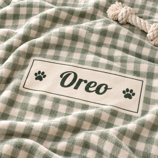 A close-up of a plush blanket with a green and cream gingham check pattern, featuring a custom white rectangular patch with the name 'Oreo' in script and two paw prints. A thick cream rope is visible in the top right corner.