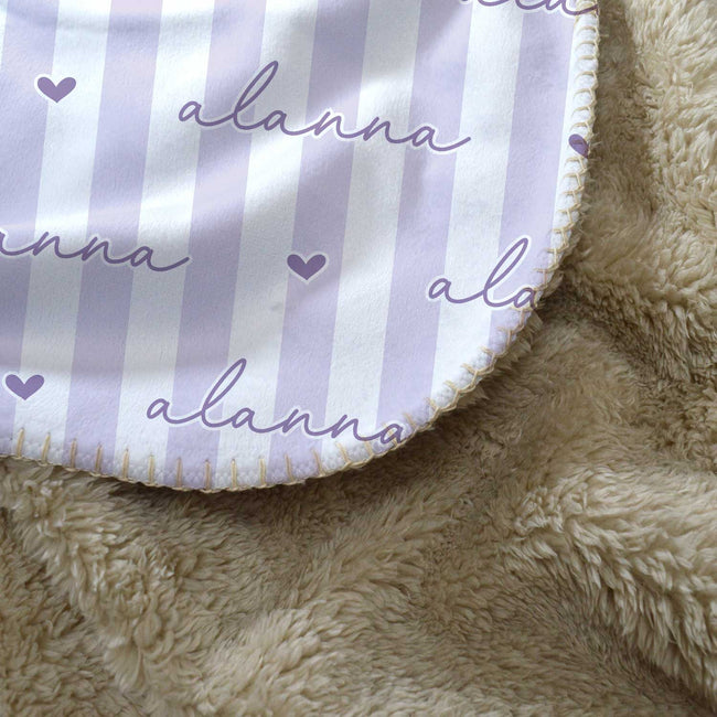 Close-up of a personalized blanket featuring lavender and white vertical stripes with the name 'alanna' and small heart motifs on one side, layered over a soft beige sherpa fleece.