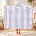 A child standing in a bedroom holding up a hooded blanket. The fabric has vertical light lavender and white stripes with a repeating pattern of the word 'alanna' in dark lavender and small lavender hearts.
