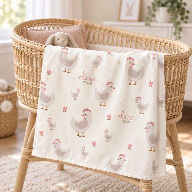 A cream-colored baby blanket with a pattern of gray chickens, pink tulips, and the text 'clara mae' is draped over a woven rattan bassinet in a bright nursery setting.