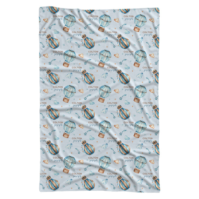A swaddle blanket featuring a repeating pattern of blue and orange striped hot air balloons, small hearts, and the text 'COLTON PYPF' on a light blue background.