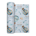 A rolled baby swaddle blanket featuring a light blue background with watercolor illustrations of striped hot air balloons, clouds, stars, and the custom name 'COLTON joseph' in brown and blue lettering.