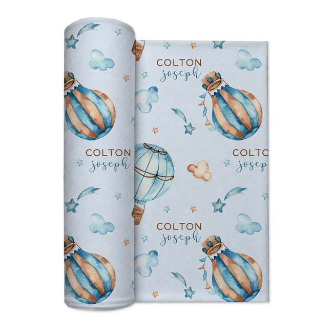 A rolled baby swaddle blanket featuring a light blue background with watercolor illustrations of striped hot air balloons, clouds, stars, and the custom name 'COLTON joseph' in brown and blue lettering.