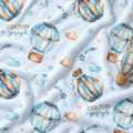 A close-up of a light blue fabric with a repeating watercolor print of hot air balloons, stars, and clouds, plus the text 'COLTON joseph' printed on it.