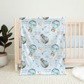 A light blue baby blanket with a watercolor print of hot air balloons, clouds, and stars, featuring the personalized names 'COLTON & joshua' in gold. The blanket is draped over a light wood crib in a nursery setting next to a potted plant.