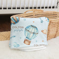 A personalized baby blanket with a light blue background, watercolor hot air balloon, stars, and the name 'COLTON joseph' printed on it, folded and draped over a woven basket.