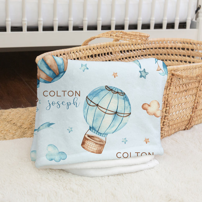 A personalized baby blanket with a light blue background, watercolor hot air balloon, stars, and the name 'COLTON joseph' printed on it, folded and draped over a woven basket.