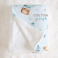 A personalized baby blanket with a white plush reverse side, featuring a light blue printed front with watercolor-style hot air balloons, stars, and the names 'COLTON joseph' in brown and blue lettering, laid flat on a white faux fur surface.
