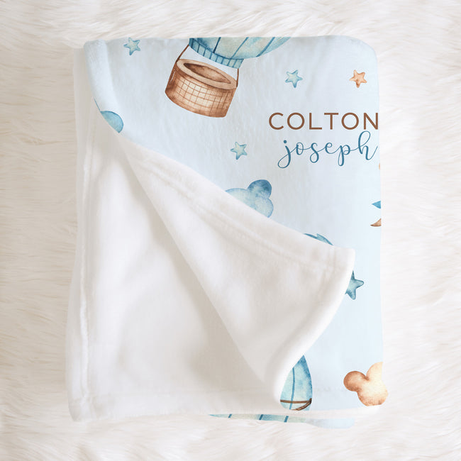 A personalized baby blanket with a white plush reverse side, featuring a light blue printed front with watercolor-style hot air balloons, stars, and the names 'COLTON joseph' in brown and blue lettering, laid flat on a white faux fur surface.
