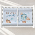 A white changing table with a personalized changing pad cover featuring a light blue fitted sheet with watercolor illustrations of a hot air balloon, clouds, mountains, and stars, along with the names 'COLTON joseph' in brown and blue text.