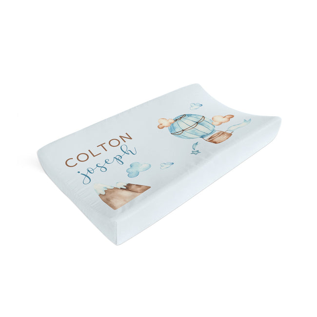 A personalized changing pad cover in white with a watercolor hot air balloon design and the names 'COLTON joseph' printed on the front, shown on a clean white background.