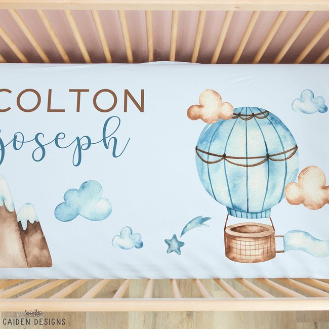 Personalized baby bedding set in a wooden crib, featuring a light blue duvet cover with watercolor illustrations of a hot air balloon, clouds, mountains, and stars, along with the names 'COLTON Joseph' in brown and blue text.