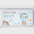 Personalized baby bedding set in a white crib, featuring a light blue background with watercolor illustrations of a hot air balloon, clouds, mountains, and stars, with the names 'COLTON' and 'Joseph' printed on the front.