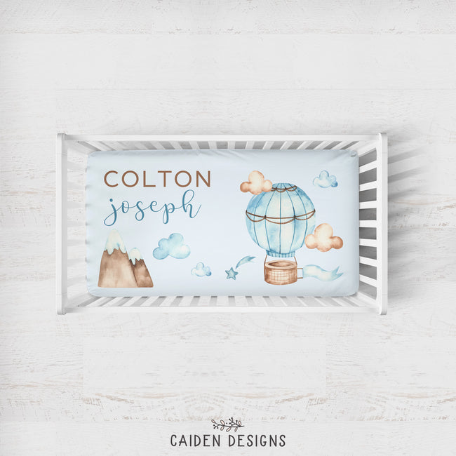 A personalized baby bedding set displayed in a white wooden crib, featuring a printed bassinet cover with watercolor illustrations of a hot air balloon, clouds, mountains, and the names 'COLTON joseph'.