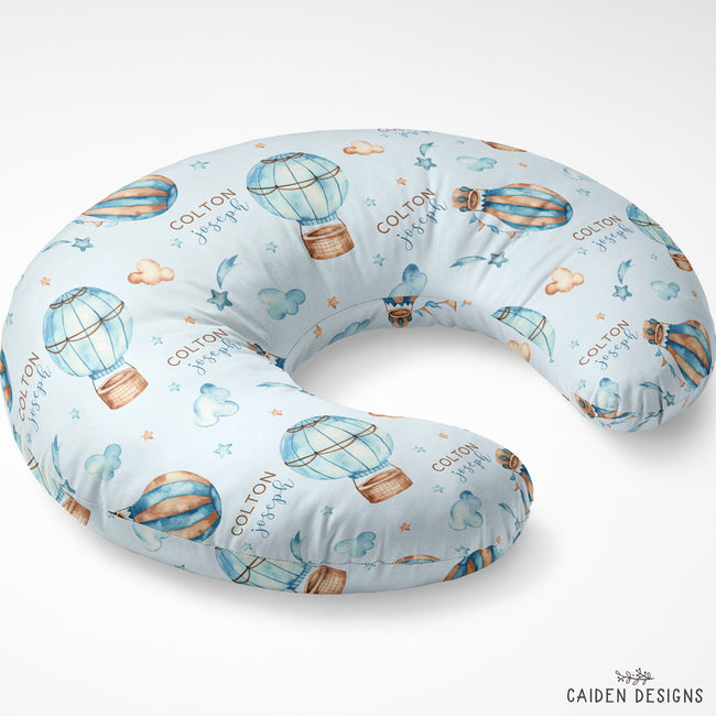 A U-shaped baby nursing pillow with a light blue background and an all-over print of hot air balloons, clouds, stars, and the name 'Colton' in script. The brand 'CAIDEN DESIGNS' is visible in the bottom right corner.