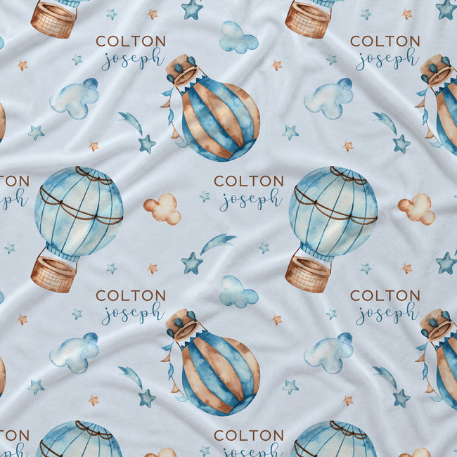 Light blue fabric with a repeating pattern of watercolor hot air balloons, stars, clouds, and the personalized name 'COLTON joseph' in brown and blue lettering.