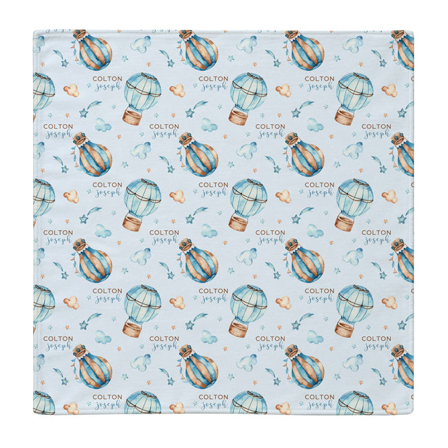 A square personalized swaddle blanket featuring a repeating pattern of blue and gold hot air balloons, small hearts, stars, and the text 'COLTON & joseph' on a pale blue background.