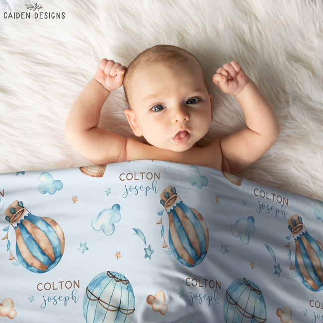 A baby lying on a white faux fur blanket, wearing a personalized blue and gold hot air balloon blanket printed with the name 'COLTON joseph'.