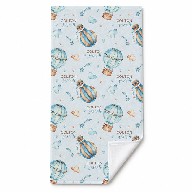 A personalized beach towel with a light blue background, featuring a repeating pattern of watercolor hot air balloons, stars, hearts, and the text 'COLTON joseph'. The blanket is shown folded to reveal the white reverse side.