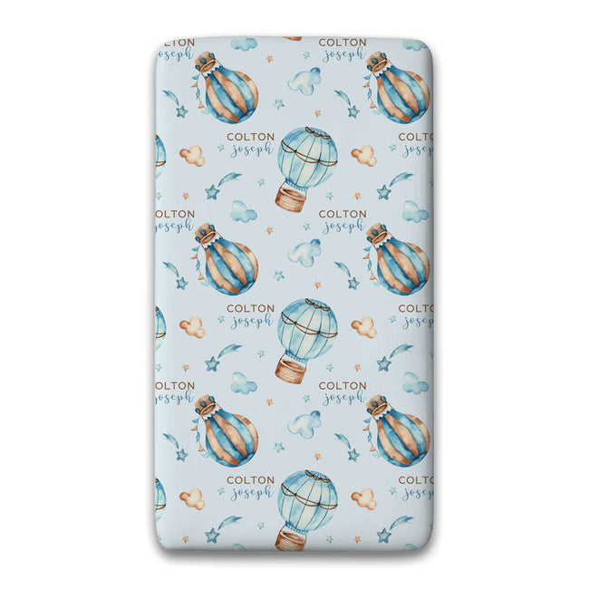 A twin fitted sheet, featuring a light blue background with a repeating pattern of watercolor hot air balloons, stars, clouds, and the personalized name 'COLTON joseph' in brown and blue.