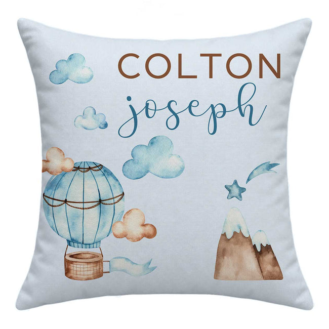 A square personalized throw pillow in light blue-gray fabric, featuring watercolor-style illustrations of a hot air balloon, clouds, mountains, and stars, with the names 'COLTON' and 'joseph' printed prominently on the front.