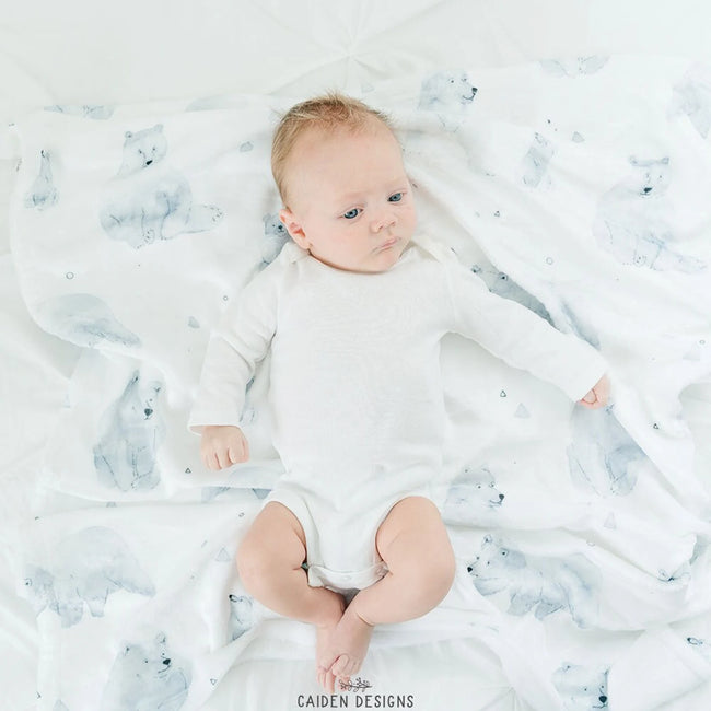A baby lying on a white blanket with a grey polar bear pattern, wearing a white long-sleeve bodysuit. The brand name 'CAIDEN DESIGNS' is printed at the bottom.