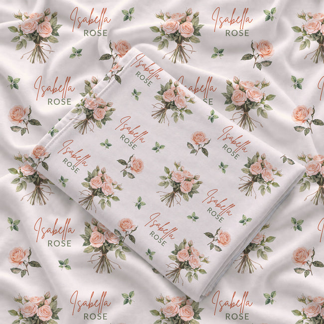 Ivory swaddle blanket with peach rose bouquets, scattered green leaves, and the name Isabella Rose in coral and green lettering.