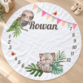 A round white baby blanketa light wood floor, featuring a gray sloth at the top, a gray baby bird at the bottom, green leaves, colorful bunting, the name 'Rowan' in the center, and numbers 1 through 12 around the edge. A woven basket and cream slippers are visible nearby.