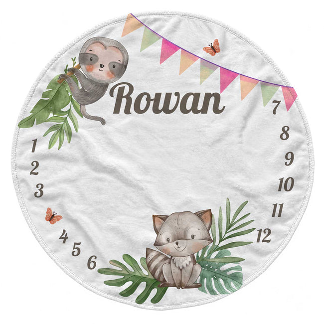 A personalized round white baby blanket featuring cute cartoon illustrations of a sloth and a baby fox, green leaves, colorful bunting, butterflies, and numbers 1 through 12 arranged around the edge. The name 'Rowan' is printed in the center.