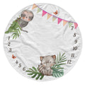 A round white baby blanket featuring a cute sloth and a baby toucan illustration, decorated with colorful pennant banners, butterflies, green leaves, and numbers 1 through 12 arranged around the border.