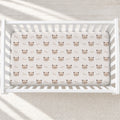 A white wooden crib with a fitted sheet featuring a repeating pattern of brown cat faces and the word 'love' in cursive, shown in a softly lit nursery setting.
