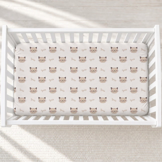 A white wooden crib with a fitted sheet featuring a repeating pattern of brown cat faces and the word 'love' in cursive, shown in a softly lit nursery setting.