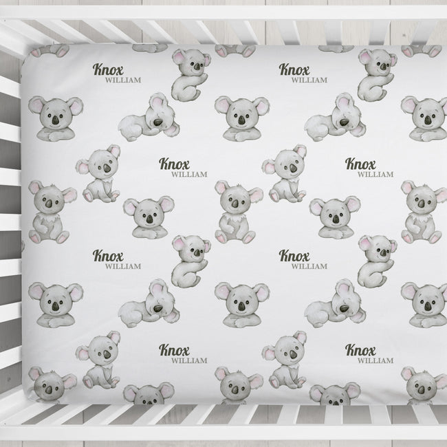 A personalized baby crib sheet with a repeating pattern of cute gray koala illustrations and names like 'Knox William', shown inside a white wooden crib.