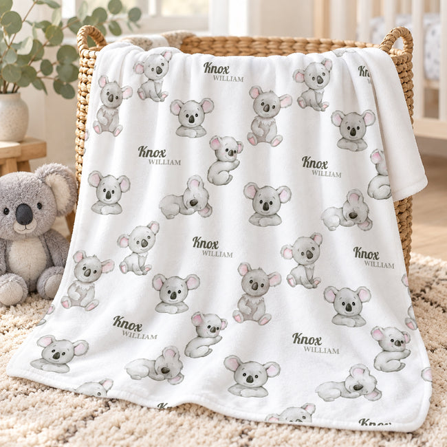 A white blanket with a repeating pattern of gray koala illustrations and the names 'Knox William' is draped over a wicker basket in a cozy room setting with a plush koala toy nearby.