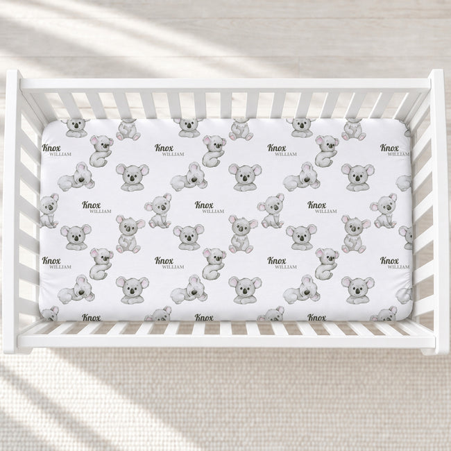 A white wooden crib with a fitted sheet printed with a repeating pattern of gray koala illustrations and the name 'Noah' in black script.