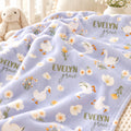 A light lavender fleece blanket with a printed pattern of white ducks, daisies, and the text 'EVELYN grace' in olive green, styled in a cozy bedroom setting with a plush bunny visible in the background.