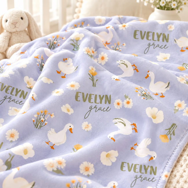 A light lavender fleece blanket with a printed pattern of white ducks, daisies, and the text 'EVELYN grace' in olive green, styled in a cozy bedroom setting with a plush bunny visible in the background.