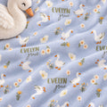 A close-up of a lavender plush blanket with a repeating pattern of white swans, small white flowers, yellow accents, and the words 'EVELYN grace' in muted green and gray. A white plush swan toy is partially visible in the top left corner.