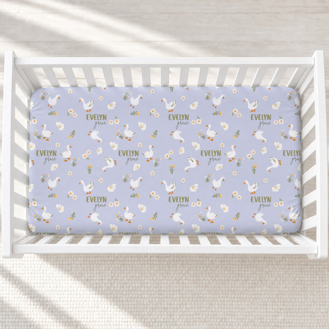 A baby crib mattress fitted sheet with a repeating pattern of white geese, daisies, and the name 'EVELYN' in sage lettering, shown inside a white wooden crib.