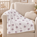 A white baby blanket draped over a beige armchair in a nursery setting, featuring a pattern of purple seashells and the name 'Riley' printed in script.