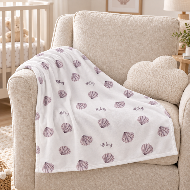 A white baby blanket draped over a beige armchair in a nursery setting, featuring a pattern of purple seashells and the name 'Riley' printed in script.
