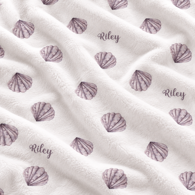 A close-up of a soft white plush blanket with a repeating pattern of purple seashells and the name 'Riley' printed in script.
