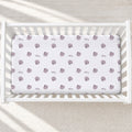 A white fitted crib sheet with a repeating pattern of mauve-purple seashells and the name 'Riley' in cursive, displayed inside a white wooden baby crib.