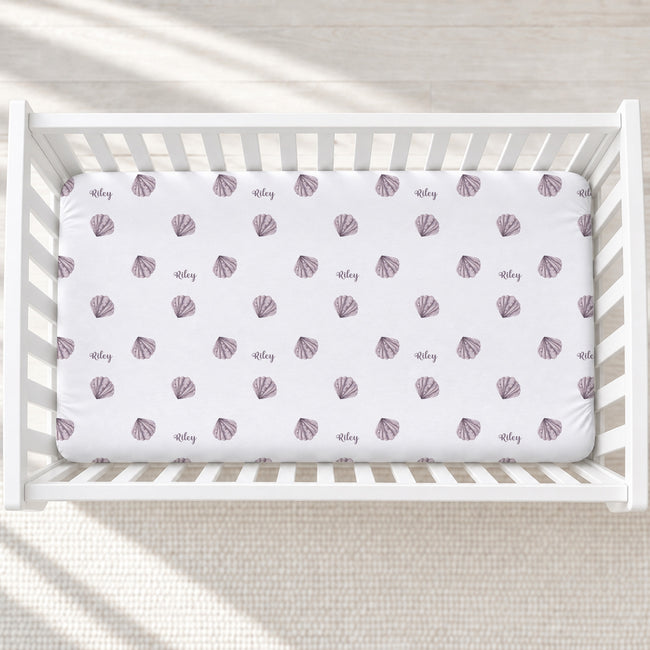 A white fitted crib sheet with a repeating pattern of mauve-purple seashells and the name 'Riley' in cursive, displayed inside a white wooden baby crib.