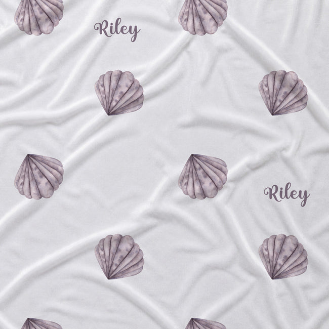 White fabric with a repeating pattern of watercolor-style lavender-gray seashells and the name 'Riley' printed in script.