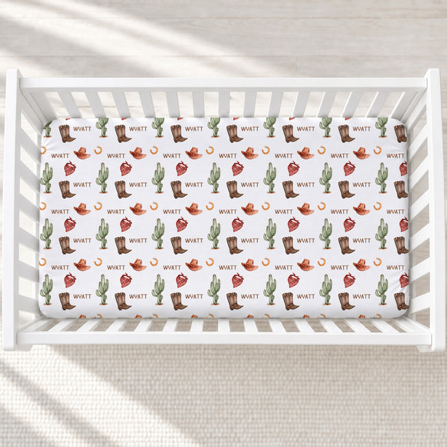 A white crib mattress fitted with a white sheet featuring a repeating western print of cacti, boots, hats, horseshoes, and the name 'WYATT' in a retro font, shown in a softly lit nursery setting.