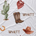 A white fabric surface with a western-themed print featuring a green cactus, a red bandana, brown cowboy boots, a brown cowboy hat, a gold horseshoe, and the name 'WYATT' printed in brown.