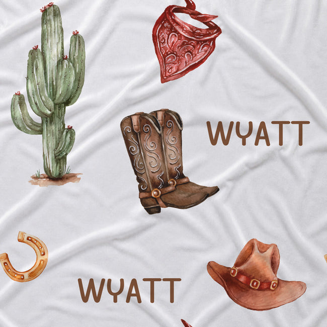 A white fabric surface with a western-themed print featuring a green cactus, a red bandana, brown cowboy boots, a brown cowboy hat, a gold horseshoe, and the name 'WYATT' printed in brown.
